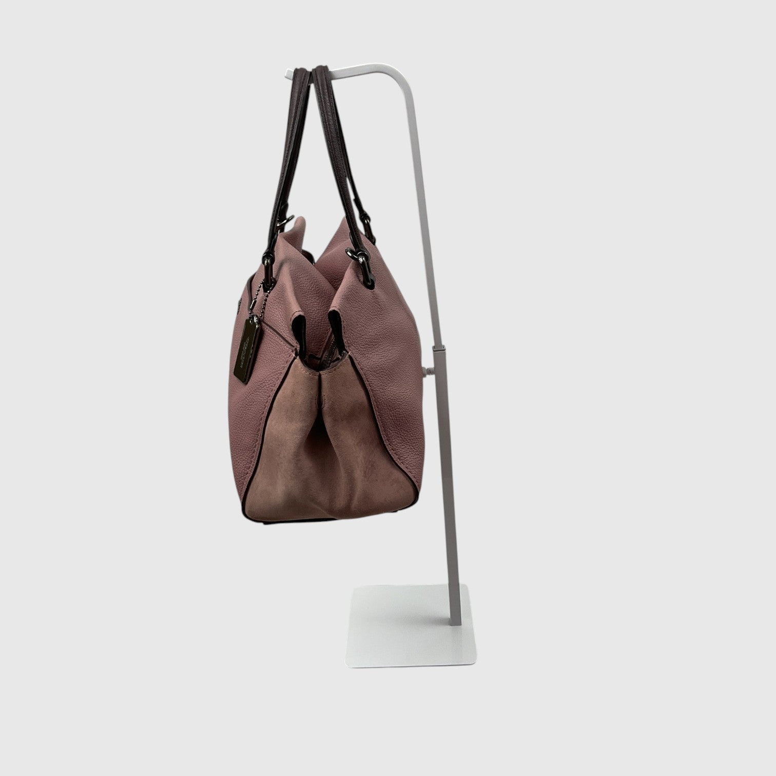 A dusty rose leather shoulder bag with black trim and straps, hanging from a minimalist white metal display stand against a light gray background.