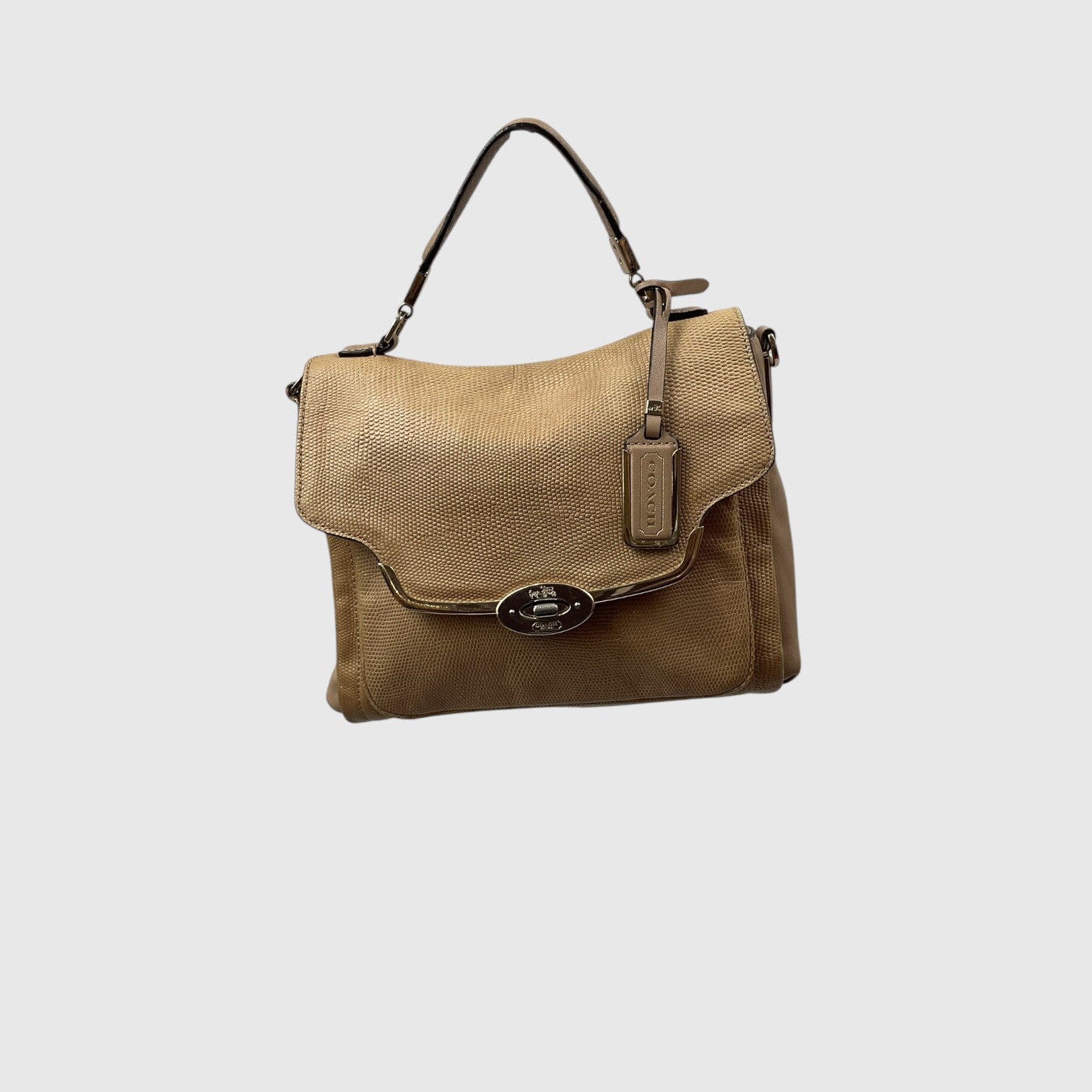 A tan textured leather handbag with a top handle, a front flap, and a silver-tone oval logo clasp, shown against a light gray background.