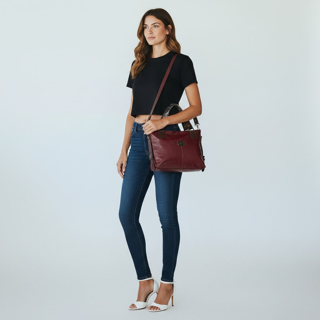 A woman modeling a dark red leather crossbody bag with a brown strap, styled with a black crop top, blue skinny jeans, and white heeled sandals against a light gray studio background.