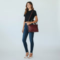 A woman modeling a dark red leather crossbody bag with a brown strap, styled with a black crop top, blue skinny jeans, and white heeled sandals against a light gray studio background.