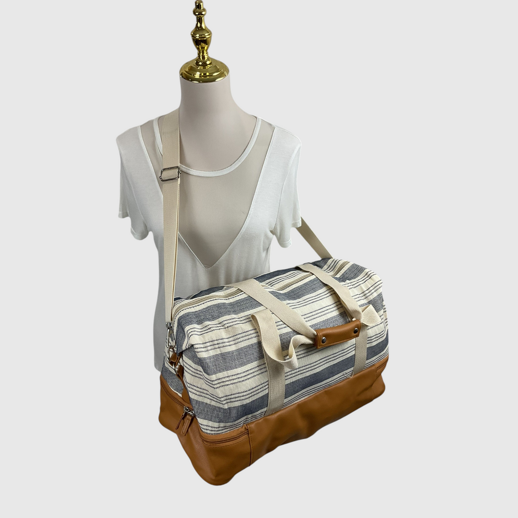 A striped canvas duffel bag with tan leather trim and a beige shoulder strap, displayed on a mannequin torso against a light gray background.