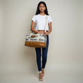 A woman modeling a large duffel bag with a cream and gray striped pattern and brown leather trim, standing against a neutral beige studio background.