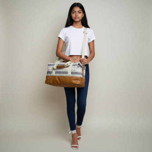 A woman modeling a large duffel bag with a cream and gray striped pattern and brown leather trim, standing against a neutral beige studio background.