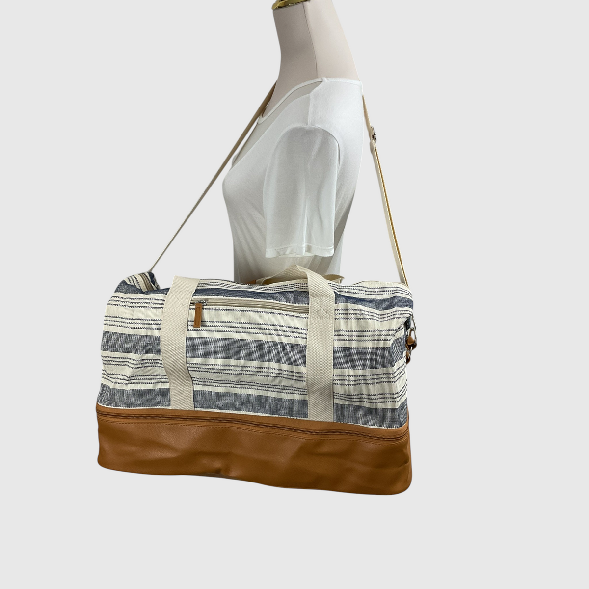 A striped canvas duffel bag with a brown faux-leather base and a tan shoulder strap, displayed on a cream mannequin torso against a light gray background.
