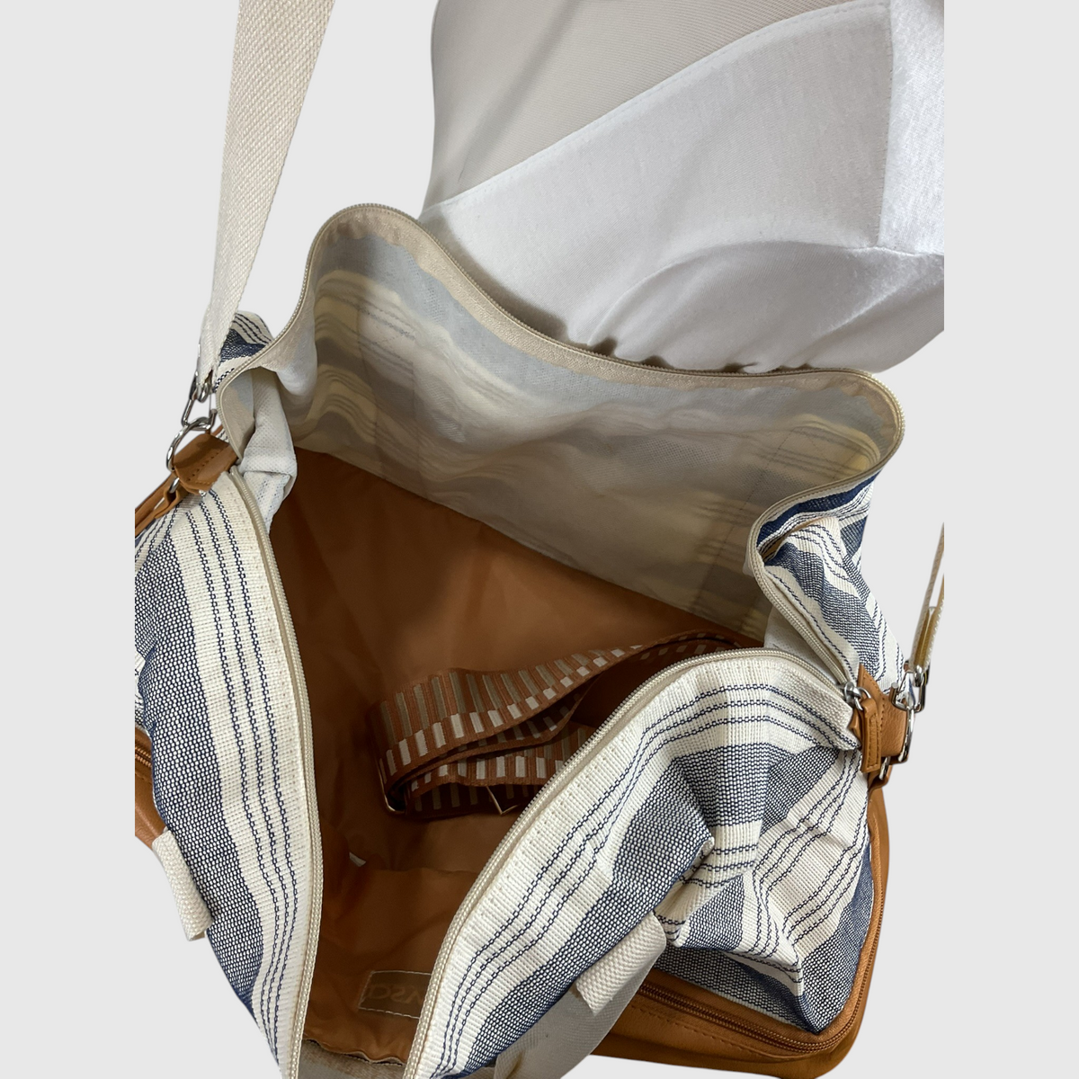 Open view of a shoulder bag with a blue and white striped woven fabric exterior, tan interior lining, and brown leather trim, shown against a light gray background.