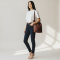 A woman stands in a bright studio wearing a white short-sleeve crop top, dark skinny jeans, white strappy heels, and carrying a large brown leather shoulder bag.