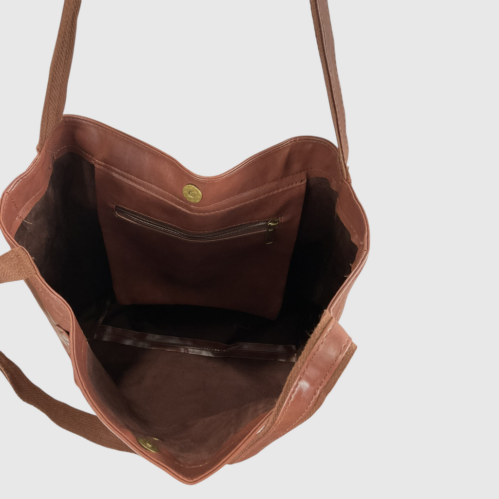 The interior of a brown leather tote bag, showing a zippered pocket on the back wall, a main compartment, and a clear plastic zippered slip pocket on the bottom, set against a plain light gray background.