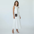 A woman modeling a sleeveless white maxi dress with a small dark brown crossbody bag and white heeled sandals, photographed against a light gray studio background.