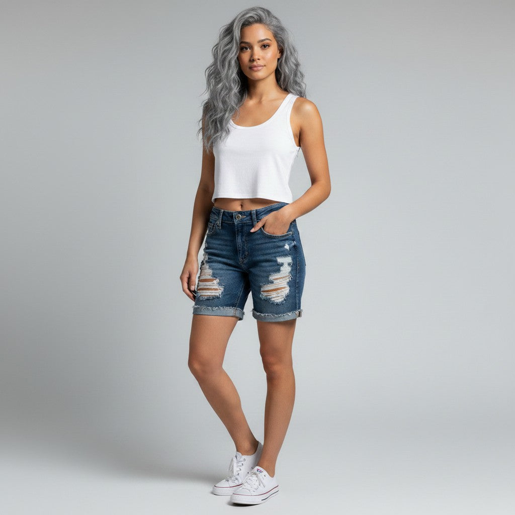 A model with silver-gray hair stands against a light gray studio background wearing a white ribbed tank top, distressed blue denim shorts with ripped details, and white sneakers.