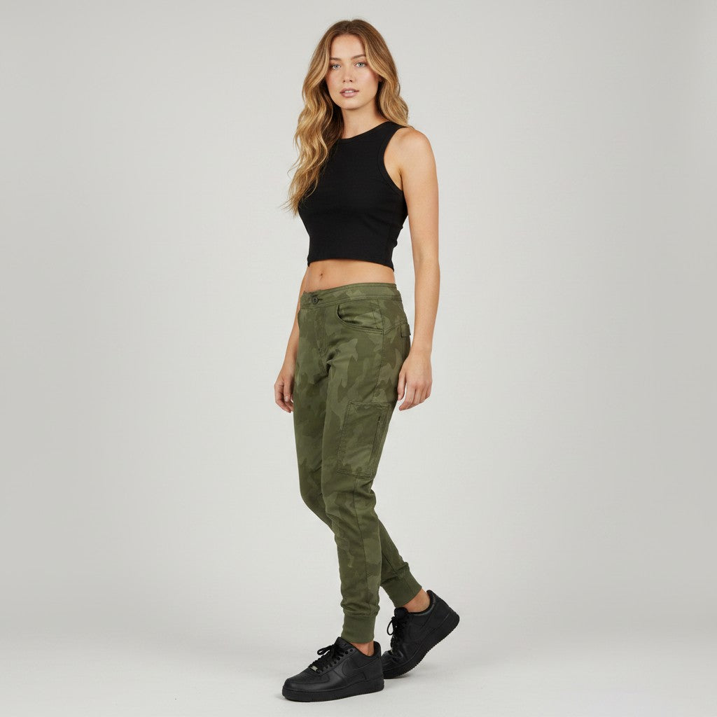 A woman modeling olive green camouflage jogger pants, paired with a black sleeveless crop top and black sneakers, against a light gray studio background.