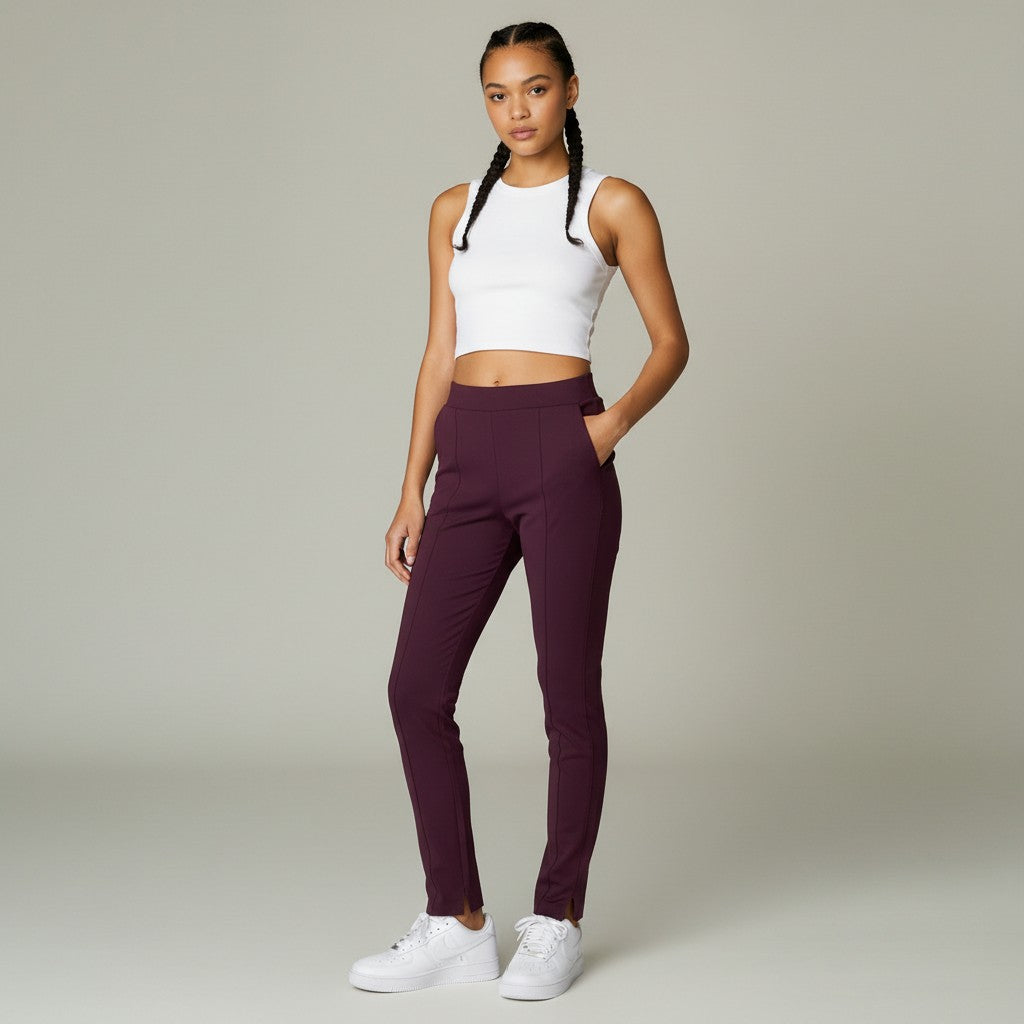 A woman modeling a pair of high-waisted, ankle-length athletic pants in deep plum purple, paired with a white crop top and white sneakers, against a solid light gray studio background.