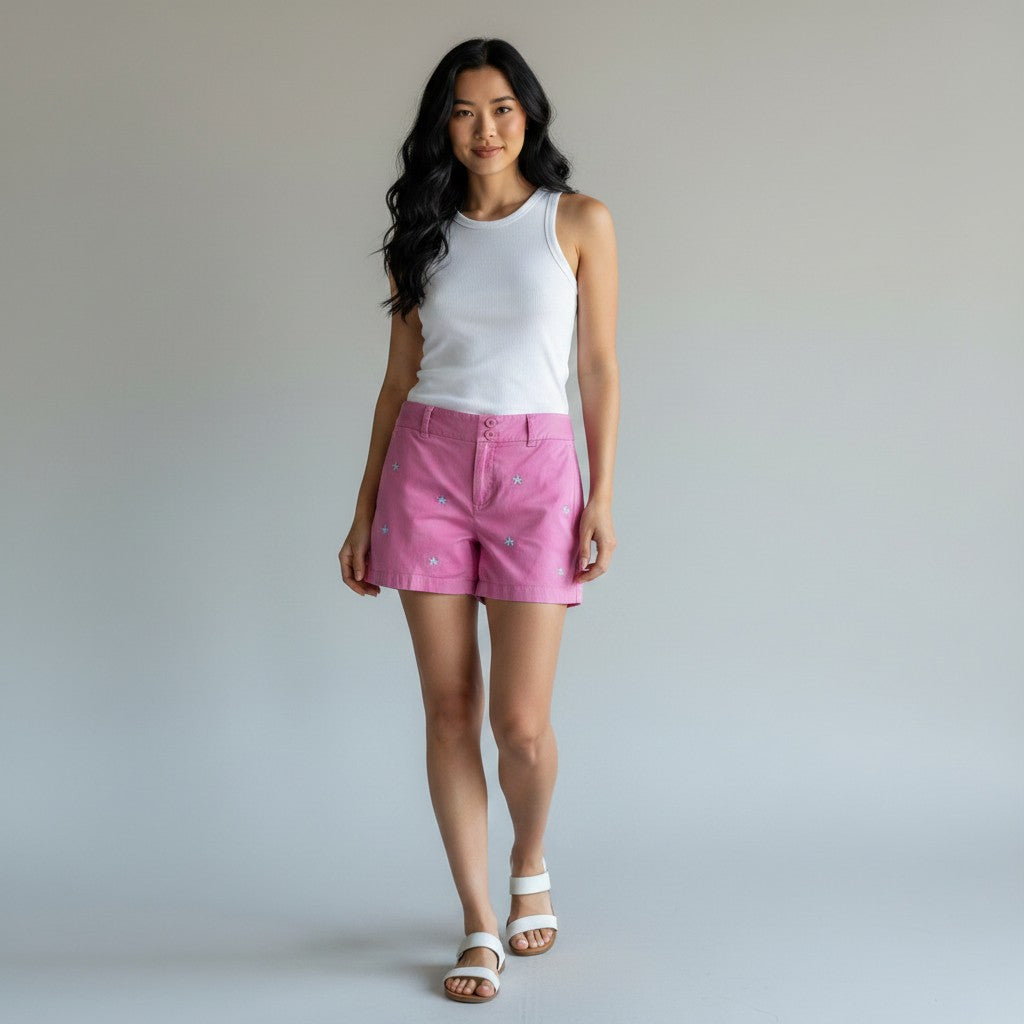 A woman wearing pink denim shorts, a white tank top, and flat sandals, standing in profile against a light gray studio background.