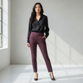 A woman stands in a bright, minimalist studio wearing a black satin long-sleeve blouse and high-waisted, ankle-length burgundy trousers with black heels.