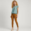 A woman modeling a light blue floral short-sleeve top and mustard-brown skinny pants with white wedge sandals, photographed against a light gray studio background.