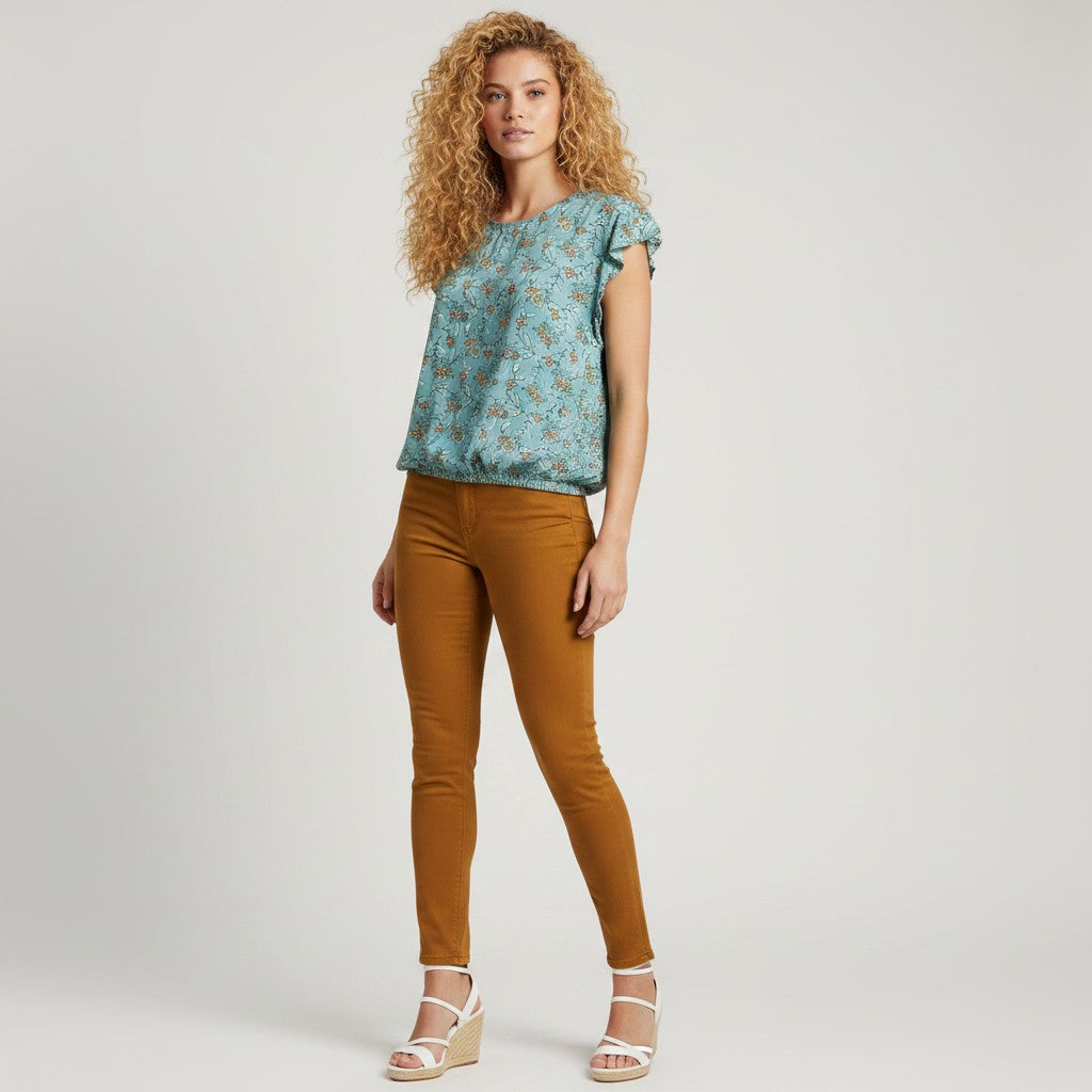 A woman modeling a light blue floral short-sleeve top and mustard-brown skinny pants with white wedge sandals, photographed against a light gray studio background.