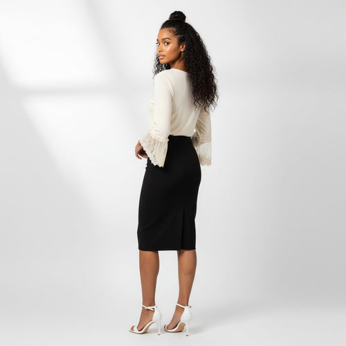 A woman stands in a studio wearing a cream long-sleeve blouse with ruffled cuffs and a black knee-length pencil skirt, paired with white high-heeled sandals. She is photographed from the back against a light gray background.