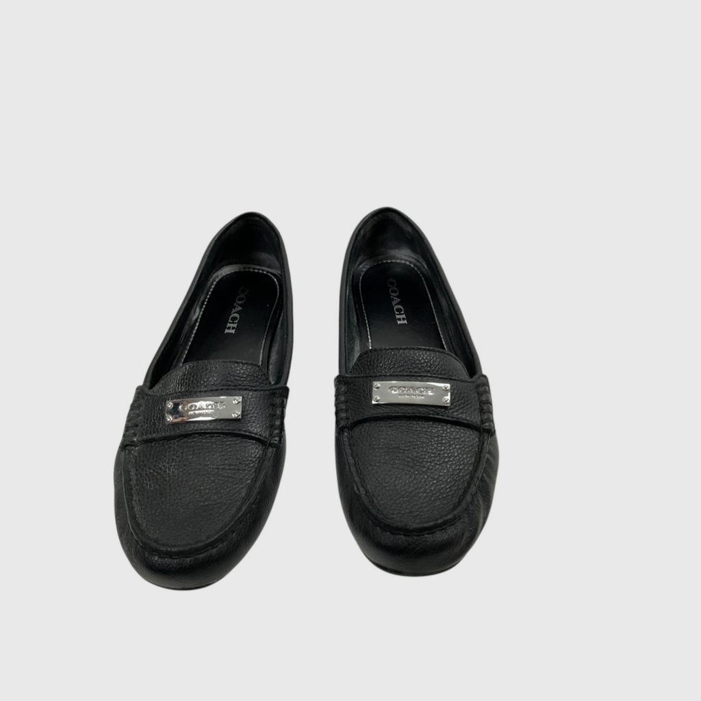 A pair of black Coach loafers with silver-tone metal logo plates on the vamp, displayed against a light gray background.