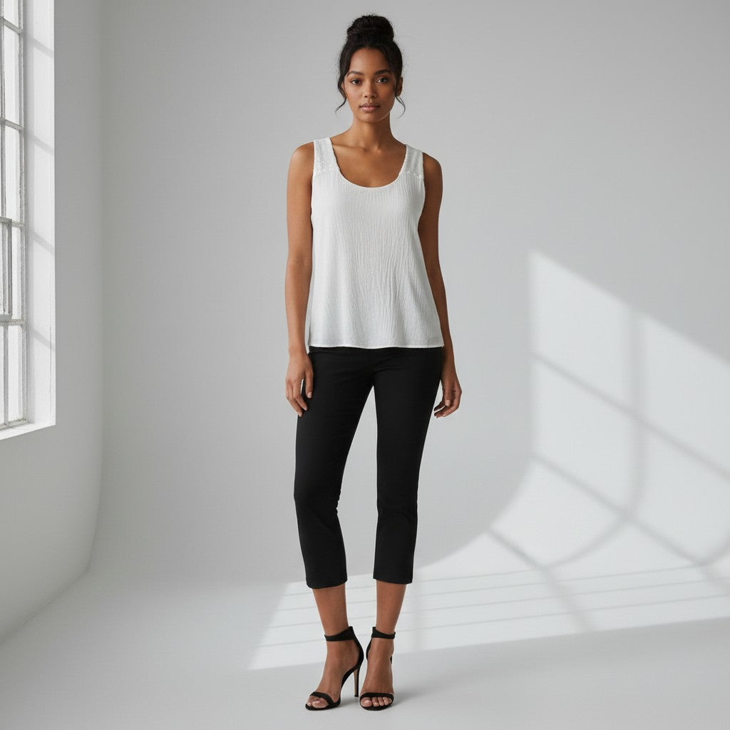 A woman stands in a bright white studio wearing a white ribbed sleeveless tank top, black cropped pants, and black heeled sandals.