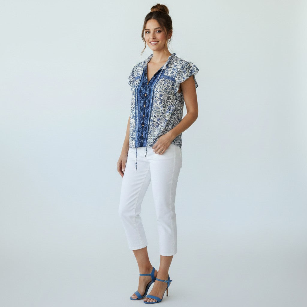 A woman modeling a short-sleeve button-front blouse with a blue and white floral pattern, paired with white cropped pants and blue high-heeled sandals, against a light gray studio background.