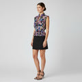 A woman models a sleeveless high-neck blouse with a colorful floral print in purple, blue, and pink, paired with a black mini skirt and black strappy heels, against a light gray studio background.