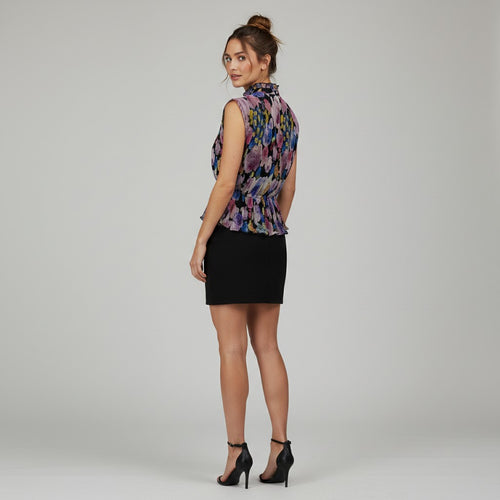 A woman modeling a sleeveless high-neck peplum top with a colorful floral pattern in purple, blue, and pink, paired with a black mini skirt and black high-heeled sandals, photographed against a light gray studio background.