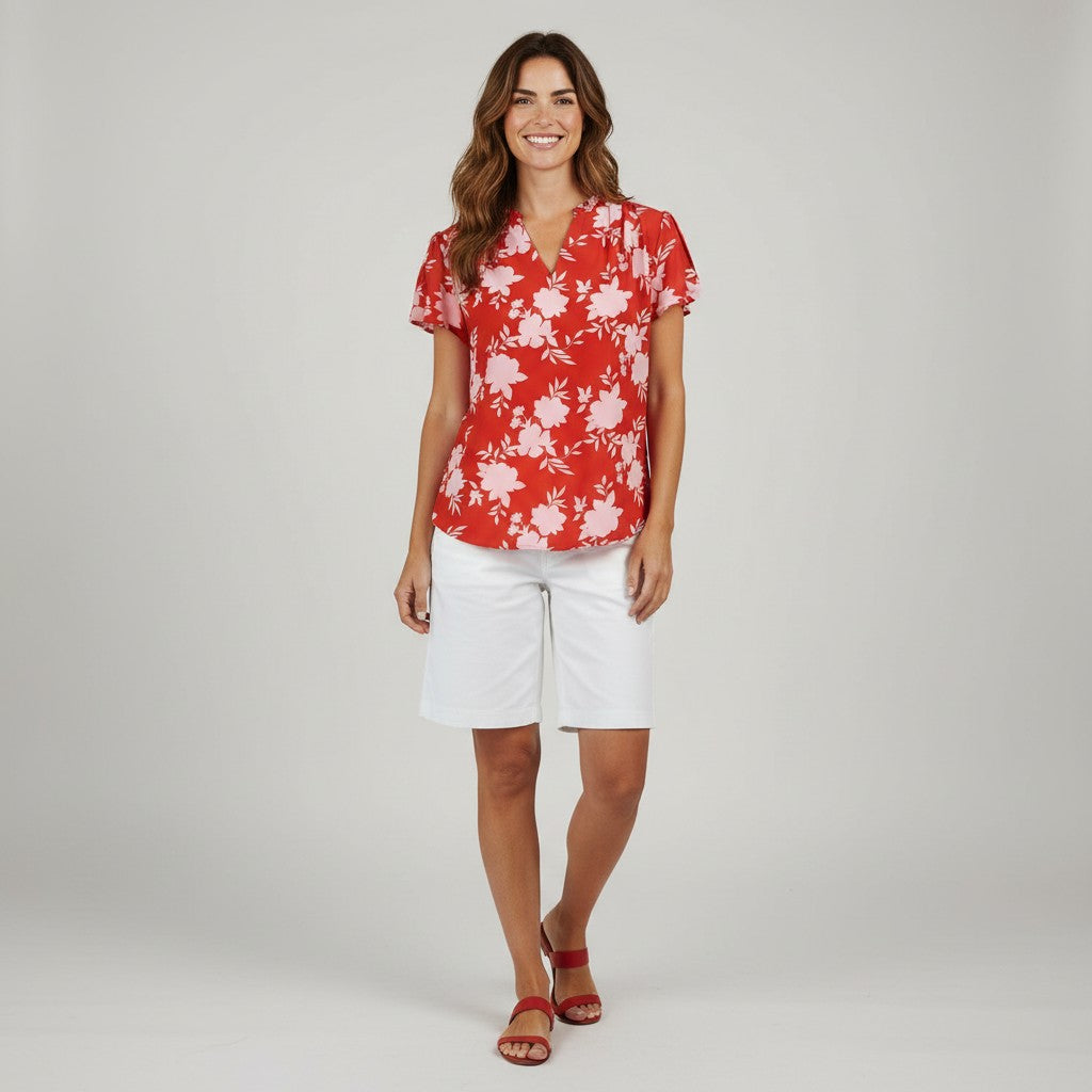A woman modeling a red short-sleeve blouse with a white floral pattern, paired with white shorts and red sandals, against a light gray studio background.