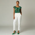 A model stands against a light gray studio background wearing a green floral-patterned short-sleeve top, white trousers, and green pointed-toe heels.