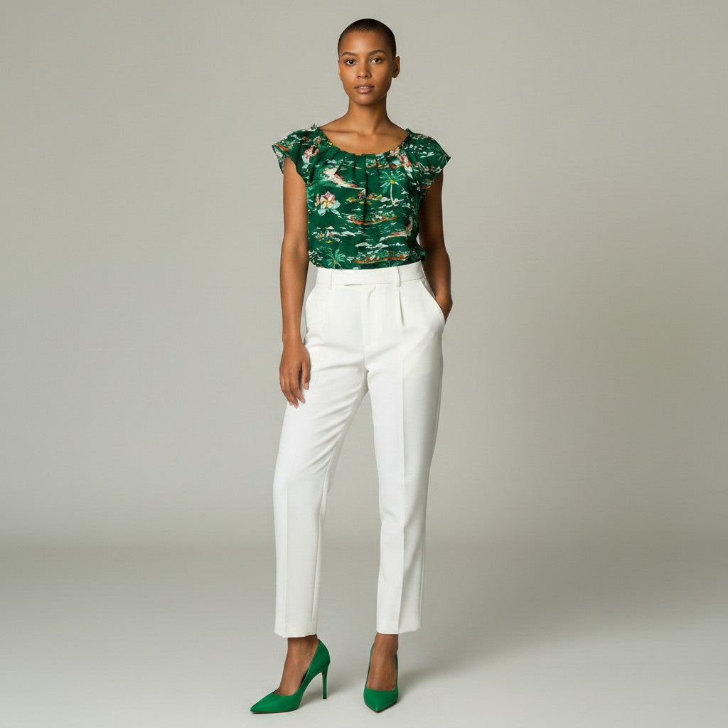 A model stands against a light gray studio background wearing a green floral-patterned short-sleeve top, white trousers, and green pointed-toe heels.