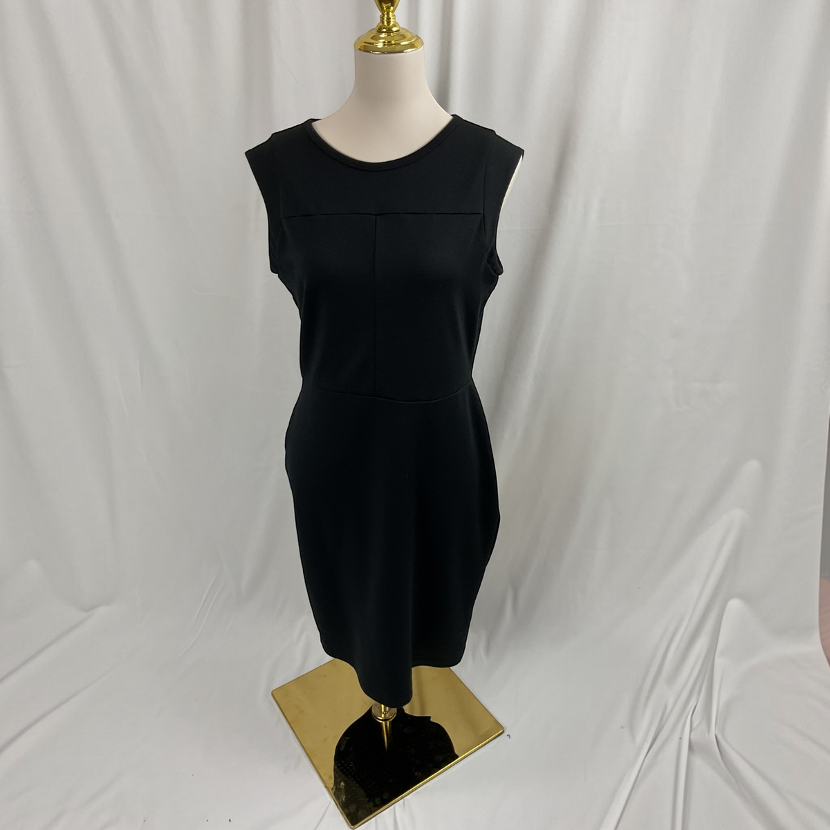 A sleeveless black dress displayed on a mannequin with a gold-tone base against a white draped fabric backdrop.