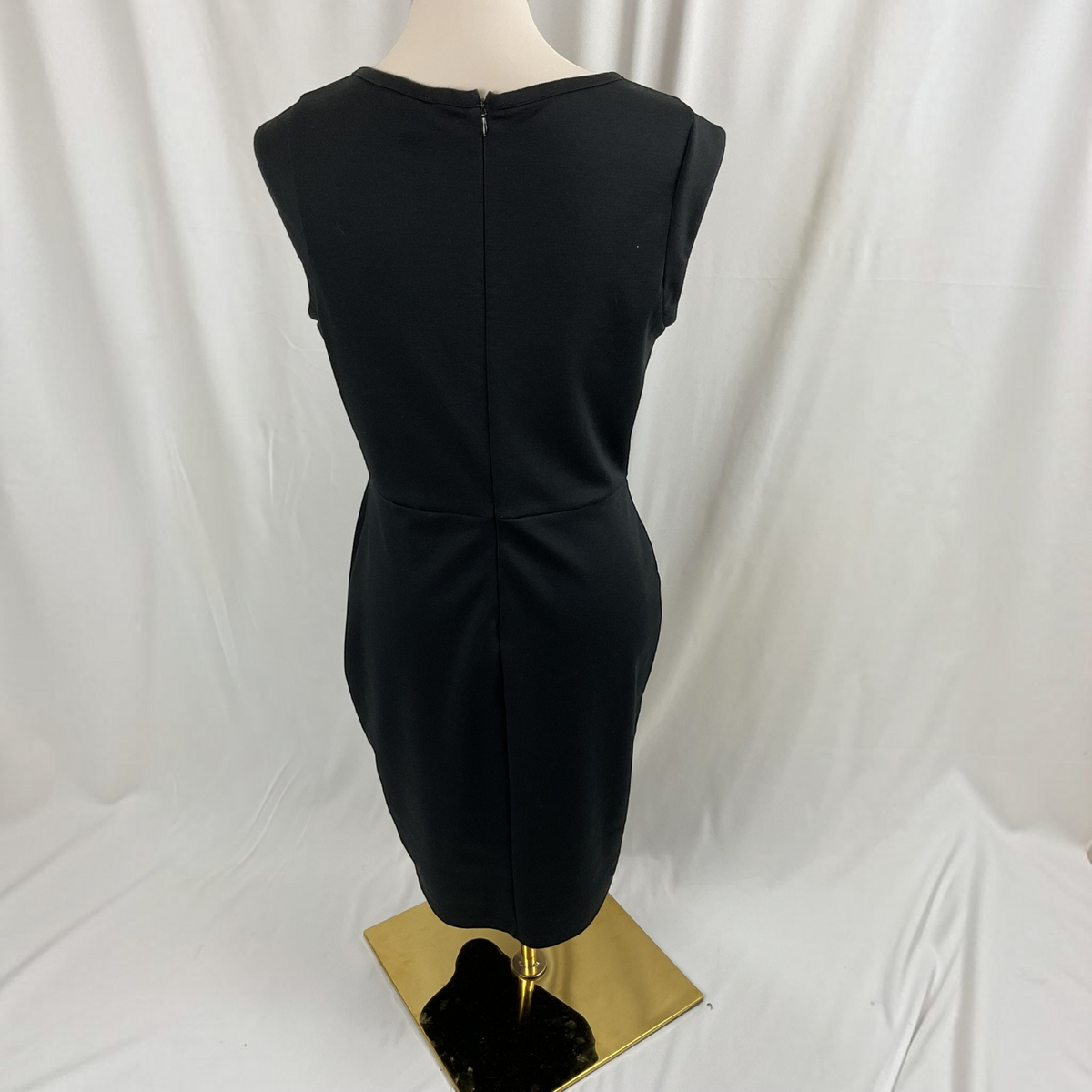A sleeveless black dress displayed on a mannequin with a gold-tone base against a white draped fabric backdrop.