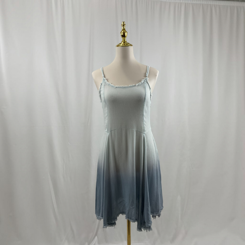 A woman modeling a sleeveless A-line dress with a light blue to slate blue ombre gradient, featuring thin spaghetti straps and an asymmetrical ruffled hem, set against a neutral studio background.