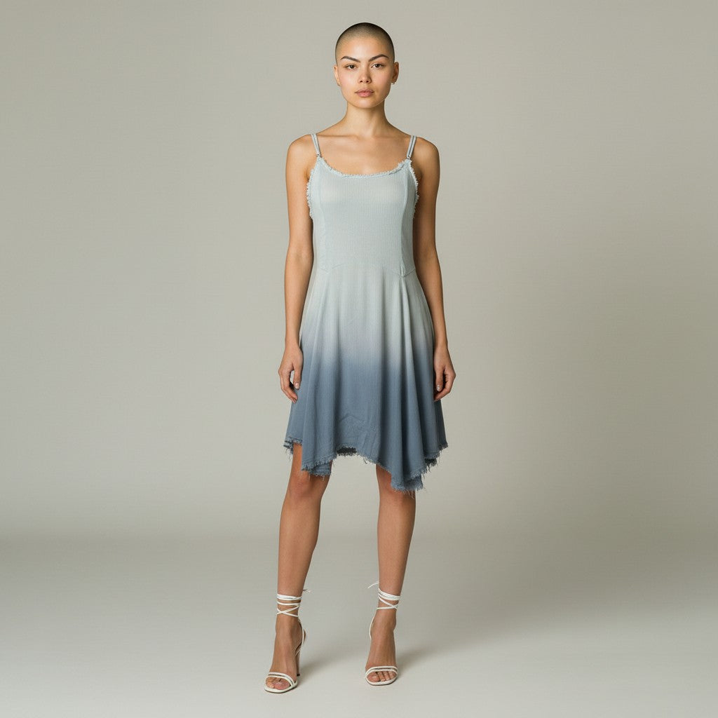 A woman modeling a sleeveless A-line dress with a light blue to slate blue ombre gradient, featuring thin spaghetti straps and an asymmetrical ruffled hem, set against a neutral studio background.