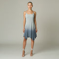 A woman modeling a sleeveless A-line dress with a light blue to slate blue ombre gradient, featuring thin spaghetti straps and an asymmetrical ruffled hem, set against a neutral studio background.