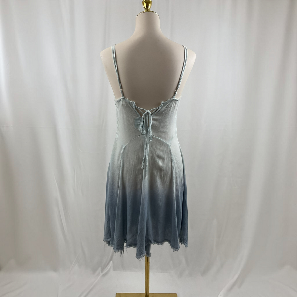 A sleeveless camisole-style dress with thin straps and a tie-front detail, displayed on a mannequin from the back against a white draped fabric backdrop. The dress has a light blue to slate blue ombré effect with a frayed hem.
