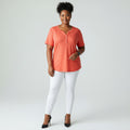 A woman with a coral shirt, white jeans, black high heels on a studio gray background.