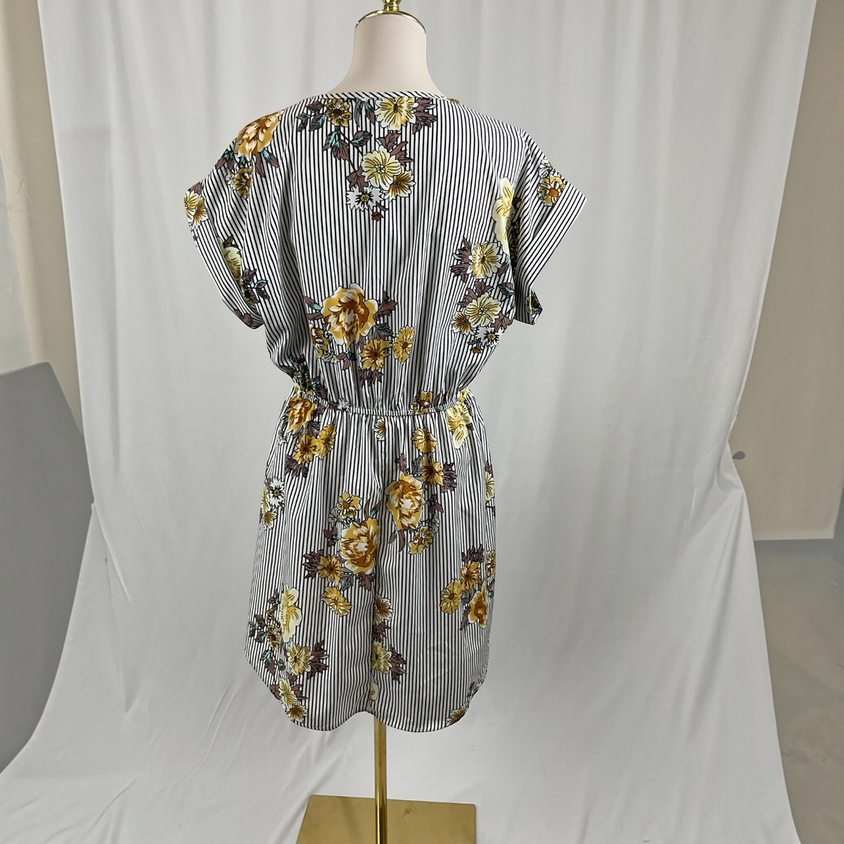 A short-sleeve V-neck dress with a gray and white vertical stripe pattern and a yellow floral print, displayed on a mannequin torso against a white draped backdrop.