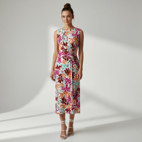 A woman stands in a studio wearing a sleeveless midi dress with a bold tropical floral print in pink, orange, brown, and light blue on a white base. She is wearing white strappy high-heeled sandals against a light gray background with soft diagonal shadows.