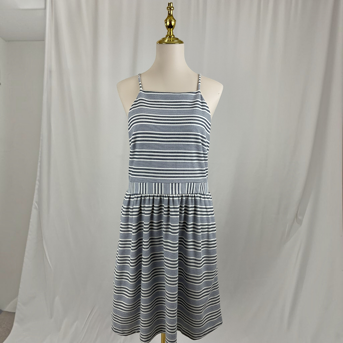 A sleeveless A-line dress with thin shoulder straps and horizontal blue, gray, and white stripes, displayed on a cream mannequin torso with a gold neck cap against a white fabric backdrop.
