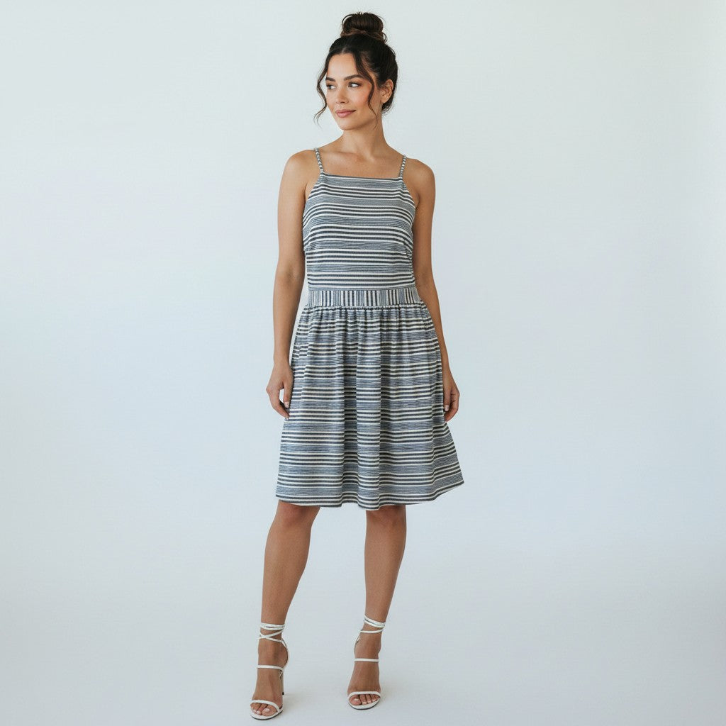 A woman modeling a sleeveless, knee-length A-line dress with thin shoulder straps and a horizontal stripe pattern in navy blue and white, paired with white lace-up heeled sandals, against a light gray studio background.