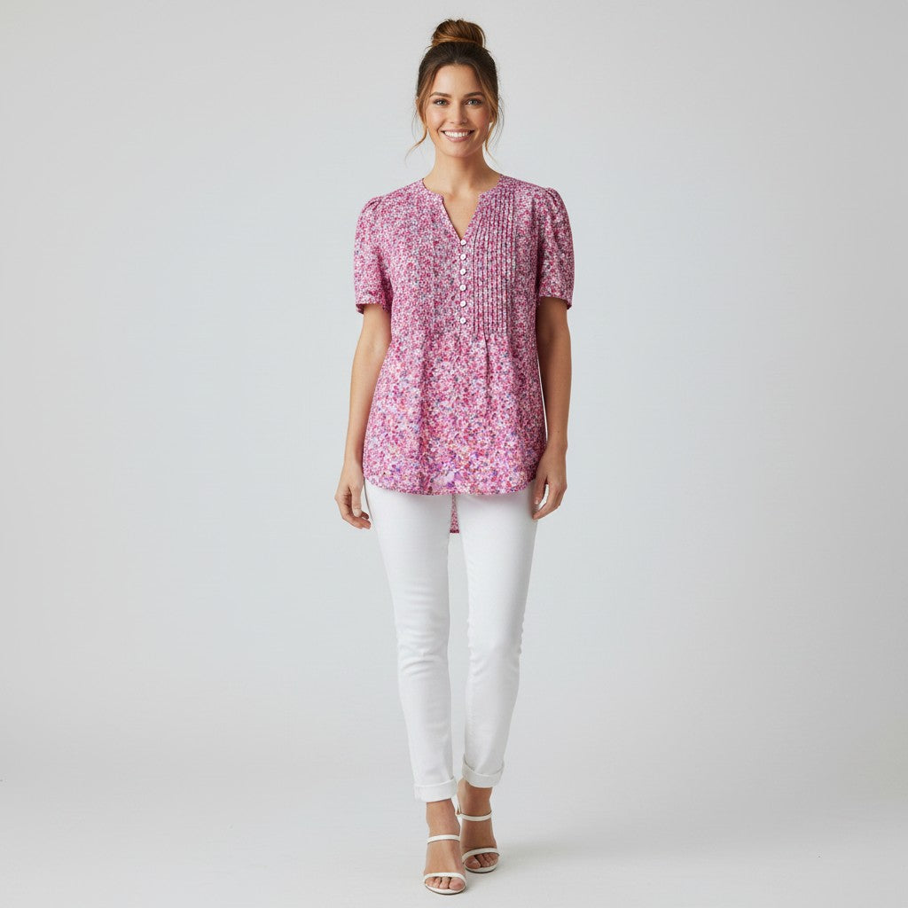 A woman modeling a short-sleeve pink and purple floral print top with a buttoned V-neck, paired with white pants and clear high-heeled sandals, against a light gray studio background.