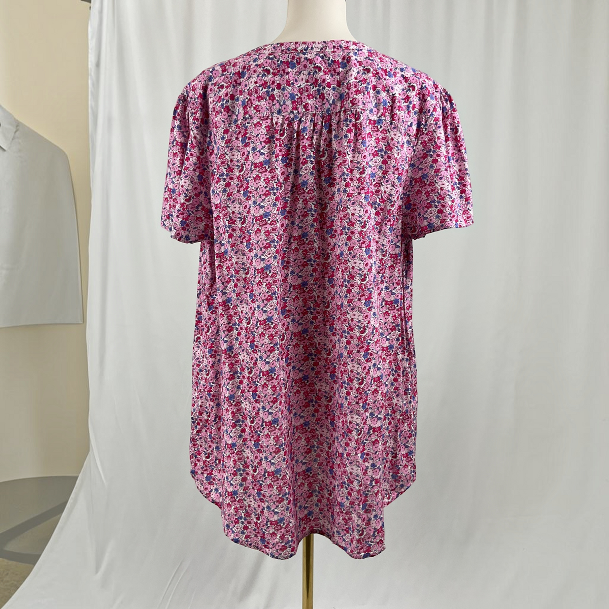 A short-sleeve blouse with a dense pink, purple, and blue floral print on a white base, displayed on a dress form mannequin against a white fabric backdrop.
