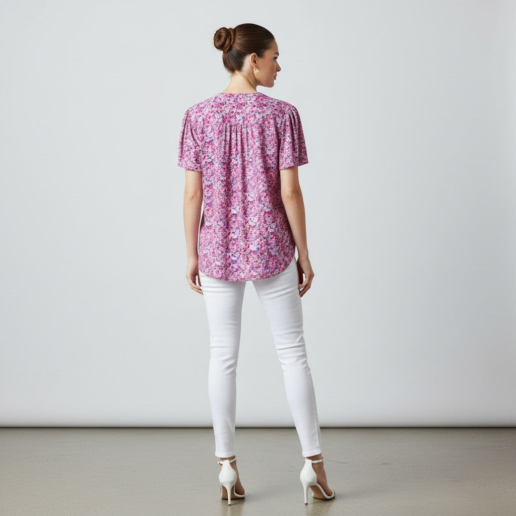 A woman viewed from the back wearing a short-sleeve pink floral top, white skinny pants, and white high heels against a plain light gray studio background.