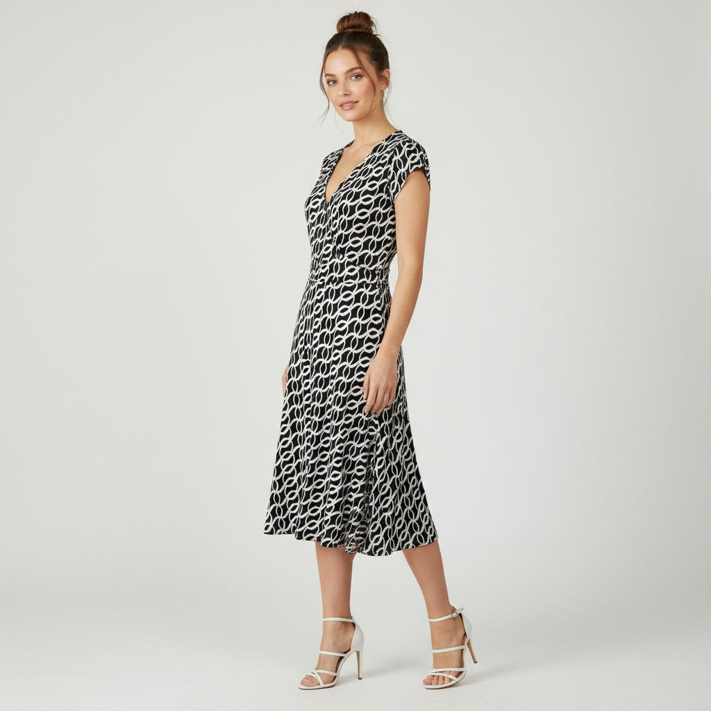 A woman modeling a sleeveless midi dress with a black and white repeating abstract pattern, photographed against a plain light gray studio background.