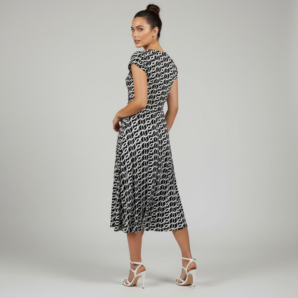 A woman modeling a sleeveless midi dress with a black and white repeating geometric pattern, shown from a three-quarters back view against a light gray studio background.