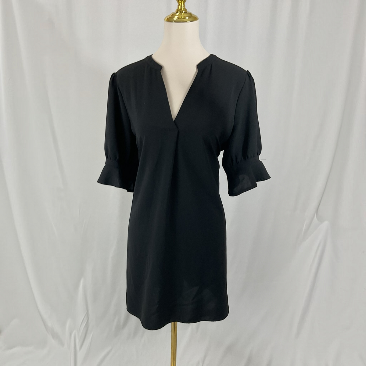 A black short-sleeve V-neck dress displayed on a mannequin with a gold stand against a white draped fabric backdrop.