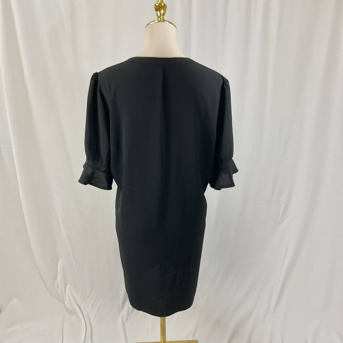 A black short-sleeve dress shown from the back on a cream dress form with a gold stand, set against a white fabric backdrop.
