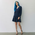 A woman with vibrant blue hair stands against a plain white wall wearing a navy blue tiered mini dress with long sleeves and brown buttons, paired with black strappy heels.