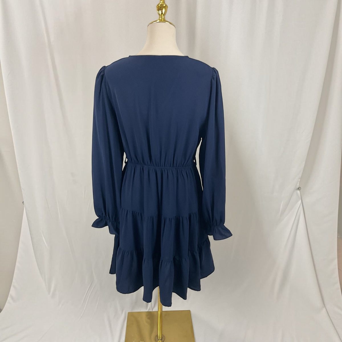 A navy blue long-sleeve dress displayed on a mannequin from the back, featuring a gathered waist and tiered skirt, set against a white draped backdrop.