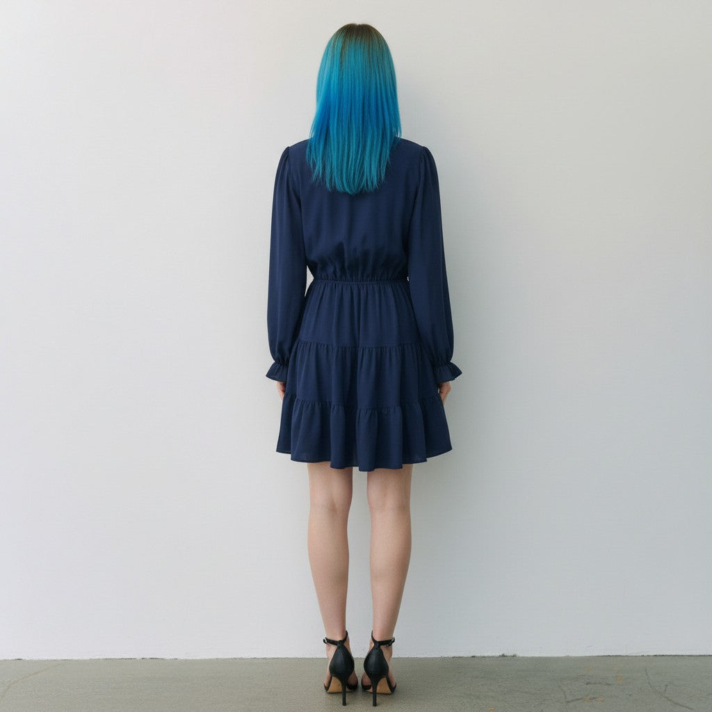 A person standing with their back to the camera wearing a navy blue long-sleeve tiered mini dress and black high-heeled sandals, set against a plain white wall.