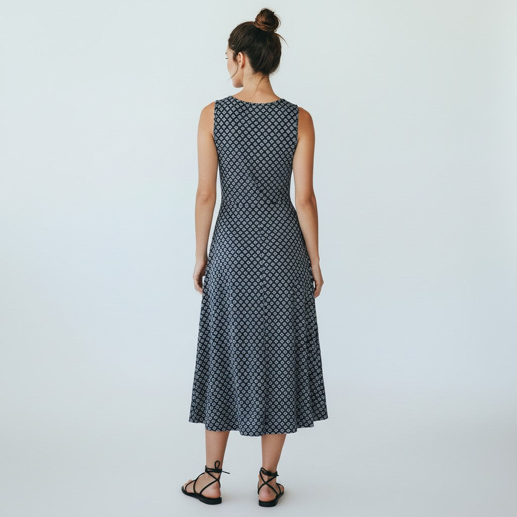 A woman viewed from behind wearing a sleeveless midi dress with a small repeating geometric pattern in dark navy and white, paired with black strappy flat sandals against a plain light gray studio background.